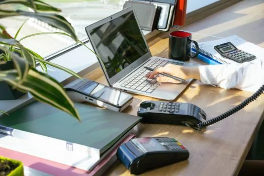 a stack of paper receipts and a calculator next to an open laptop on a desk, soft natural window light, editorial photography, shallow depth of field, no text