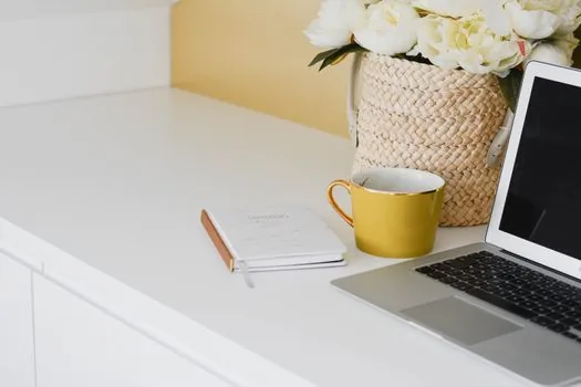 a tidy home office desk with an open laptop, a coffee mug and a small notebook by a bright window, warm morning light, editorial photography, shallow depth of field