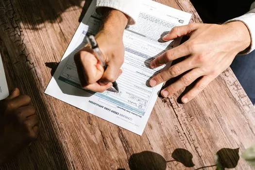 overhead close-up of hands comparing blank official notice papers with an abstract online application form, checklist tabs beside them, bright window light, clean editorial photography, no face, no logo, no readable text, no money symbols