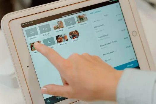 Delivery app menu management screen on tablet beside pork, kimchi, rice, seafood supplier invoices and a handwritten update checklist