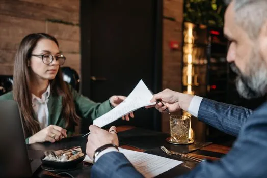 Korean cafe owner and first employee checking printed employment contract, work schedule and wage memo on wooden counter, realistic small business documentation photo | 첫 직원 채용 전 근로계약서 항목을 함께 확인하는 모습